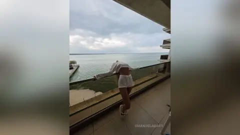 Woman in white summer outfit leaning on a balcony overlooking a calm ocean under a cloudy sky.