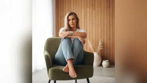 Hailey sits in a green armchair wearing a white t-shirt and blue jeans, looking directly at the camera with her bare feet visible.