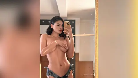Young woman with long dark hair posing in a denim bikini for a confident mirror selfie in a bright room.