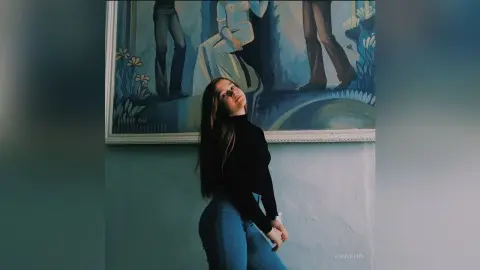 Young woman in a black turtleneck and blue jeans posing in front of a large classical painting in a moody room.