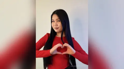 Smiling young woman in a red long-sleeve ribbed top making a heart shape with her hands against a white wall.