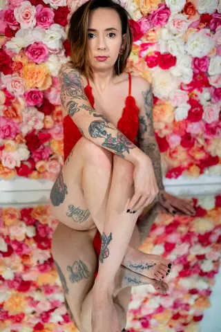 TaylaRae poses in a red lace bra against a wall of pink and white roses, showing off her tattoos and bare legs.