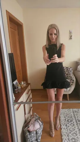 Blonde woman in a chic black sleeveless mini dress taking a mirror selfie in a bright modern bedroom.