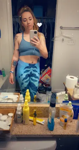 Young woman in blue yoga set taking a mirror selfie in a bathroom setting with a relaxed mood.