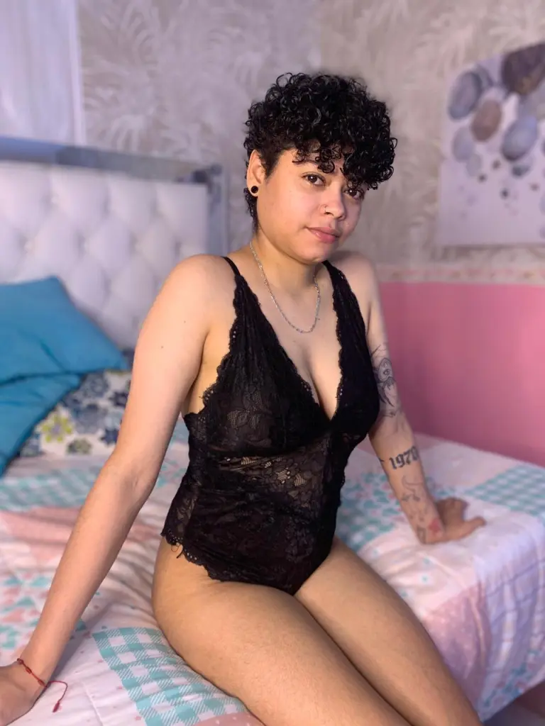 Young woman in a black lace camisole and silver necklace relaxing in a bedroom setting.