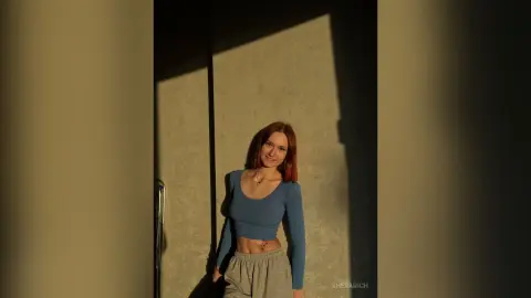 SheraRich poses in a blue long-sleeve crop top and grey sweatpants, showing her midriff and red hair against a wall with harsh shadows.