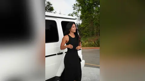 Smiling woman in a chic black sleeveless dress standing by a white van in a bright outdoor setting.