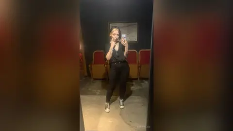 Young woman in a black shirt and pants taking a mirror selfie in a dimly lit vintage room.