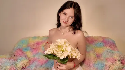 Smiling young woman with long dark hair holding white flowers against a colorful pastel faux fur background.