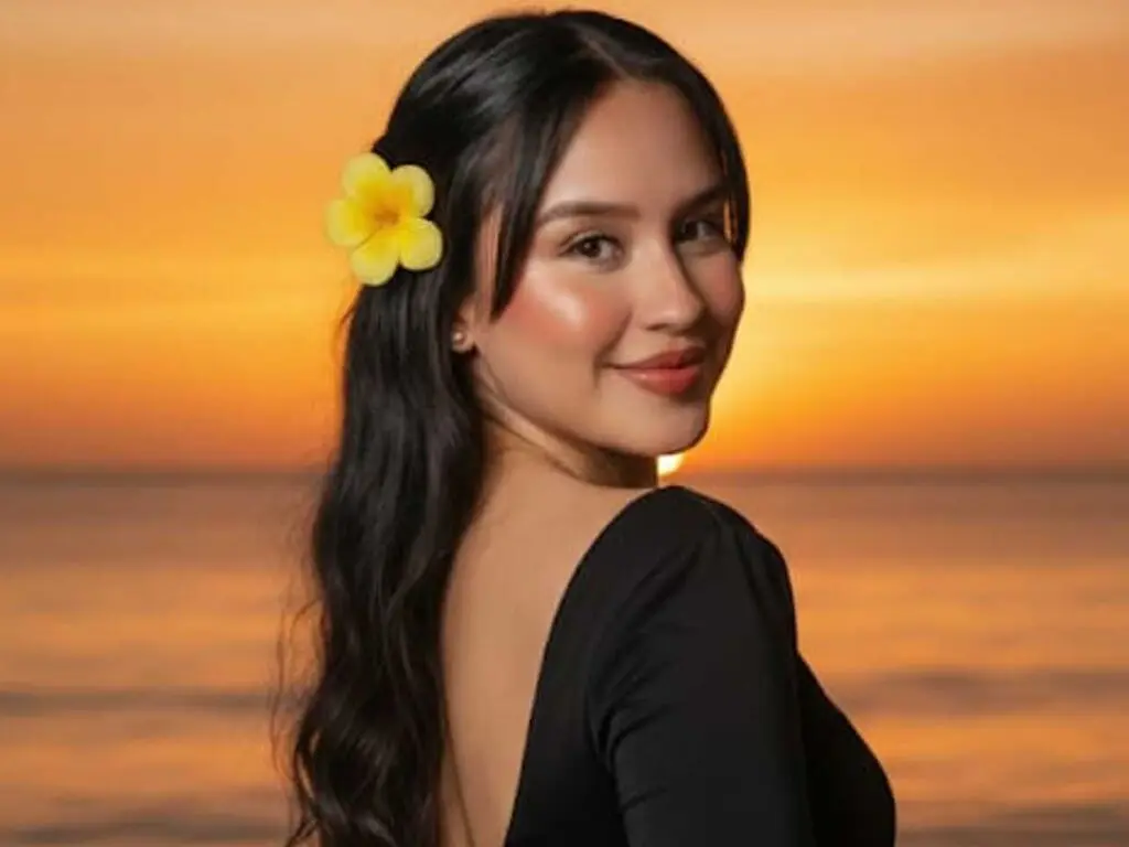 KatePolanco poses at sunset on a beach, looking over her shoulder with a yellow flower in her long dark hair and wearing a black low-cut top.