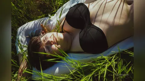 Relaxed woman in a black bikini top lying on a white towel in sunny green grass.
