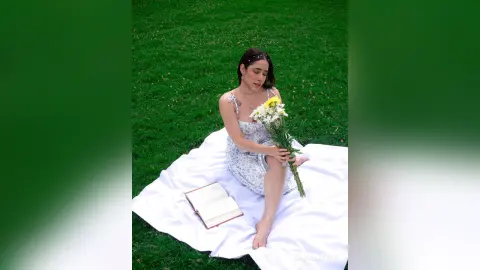 IsabellaYork — Young woman in a floral sundress sitting on a white blanket reading a book in a lush green meadow.