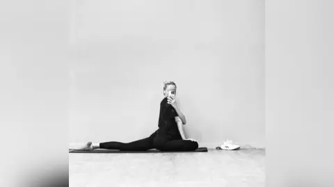 Woman in black activewear doing floor stretches on a yoga mat in a minimalist studio setting.