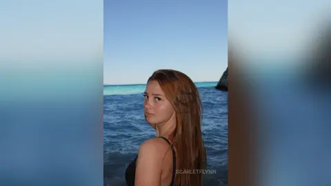 Young woman with long blonde hair in a black swimsuit relaxing by the turquoise ocean under a clear blue sky.