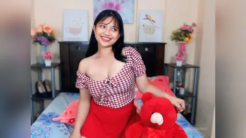 Smiling young woman in a red gingham top sitting on a bed next to a red teddy bear in a bright bedroom.