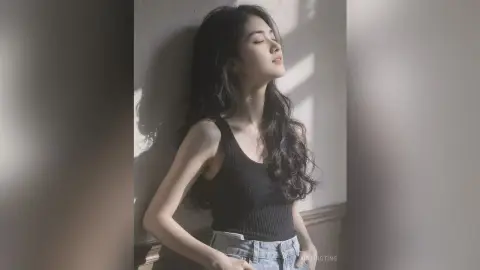 Serene young woman in a black tank top leaning against a wall in soft, dreamy sunlight.