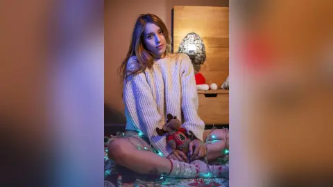 ValentinaGarcez sits cross-legged on a patterned rug wearing a white knit sweater and light-colored shorts, holding a small teddy bear.