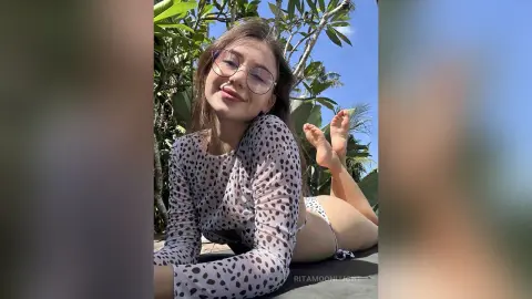 RitaMoonllight smiles while wearing a long-sleeve polka-dot top and white bikini bottoms, lying back outdoors near tropical green plants.