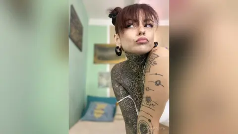 Young woman with heavy body tattoos and bangs posing in a bright room with a thoughtful expression.