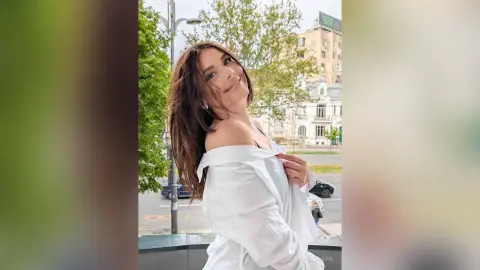 Smiling brunette woman in a white oversized shirt posing outdoors against a city street background.