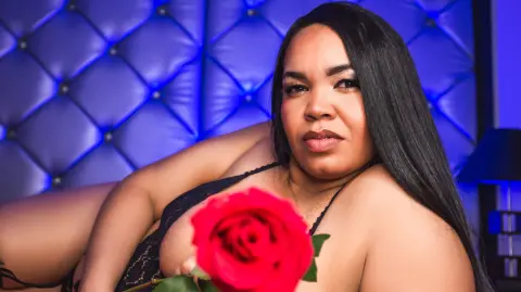 Curvy woman in black lace lingerie holding a red rose in a moody, blue-lit bedroom setting.