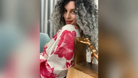 Smiling woman with curly grey hair wearing a white and red floral blouse in a bright indoor setting.