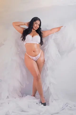 Curvy woman in white lace lingerie posing against a soft white feathered backdrop in a dreamy studio setting.
