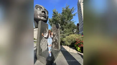 Woman in white shirt and jeans standing by a large stone face sculpture in a sunny urban park.