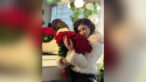 Smiling woman in a white faux fur coat holding a large bouquet of red roses in a bright room.