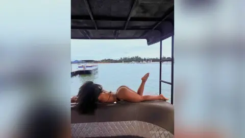 Woman in black bikini relaxing on a boat deck overlooking a calm lake on a sunny summer day.