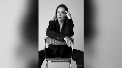 Elegant woman in a black blazer sitting on a chair in a moody black and white studio portrait.