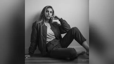 Hailey sits on the floor wearing a dark bomber jacket and matching trousers, posing with one hand near her face and long hair falling over her shoulders.