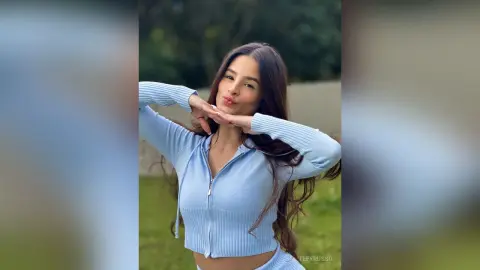 TefyRusso poses outdoors in a light blue zip-up long-sleeve crop top, framing her face with her hands and pouting her lips.