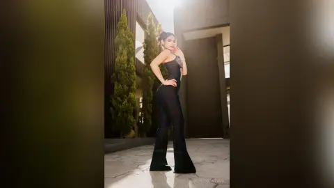 Elegant woman in a black sleeveless jumpsuit posing confidently in a modern, sunlit architectural setting.