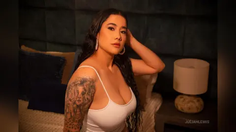 JuliahLewis poses in a white tank top, showing off a large floral tattoo on her shoulder and arm.