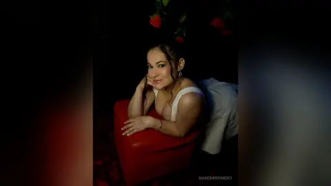NahomiRomero poses leaning over a red chair, wearing a white tank top and showing her face and shoulders against a dark background with red roses.