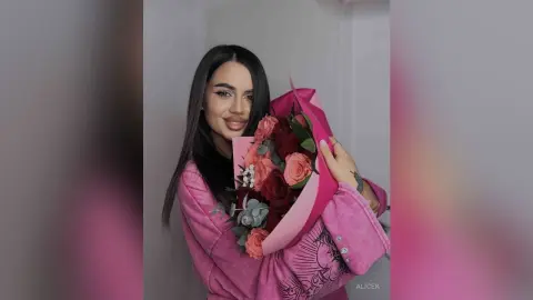 Smiling woman in a pink velvet jacket holding a large bouquet of red and peach roses in a soft indoor setting.