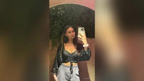 Young woman in a black long-sleeve crop top taking a mirror selfie in a moody, dimly lit room.