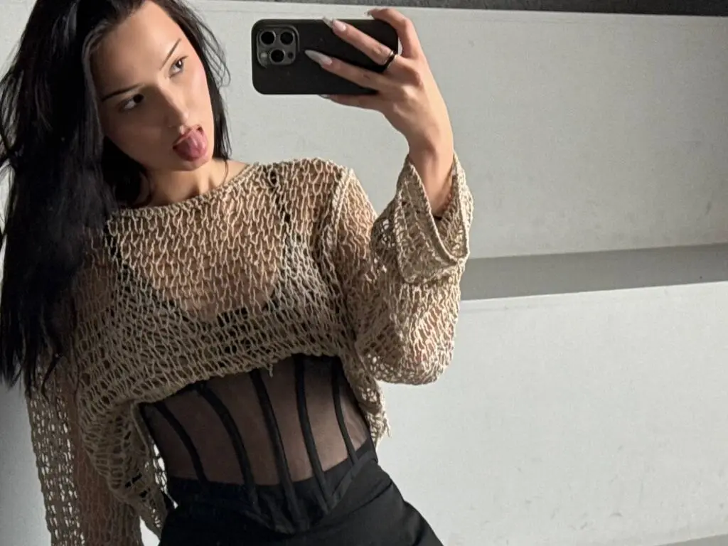 Young woman in a beige crochet mesh top and black corset bodysuit taking a moody mirror selfie.