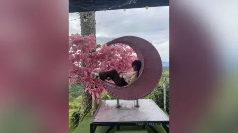 Relaxed woman in casual attire lounging in a crescent moon swing chair amidst blooming pink cherry blossoms.