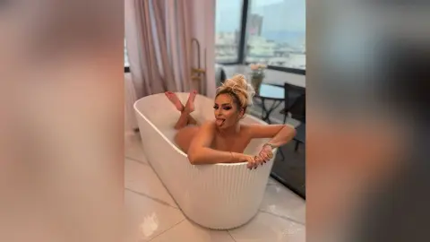 Glamorous blonde woman in a white bathtub relaxing in a modern luxury apartment with a playful mood.