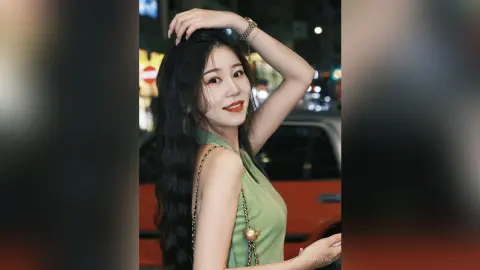 Elegant woman in a green sleeveless top posing with long wavy hair in a nighttime city street setting.