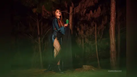 AmberNash — Mystical woman in a dark flowing gown holding a green lantern in a moody, foggy forest at night.