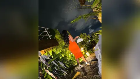 Elegant woman in an orange midi dress walking through a tropical garden at night.