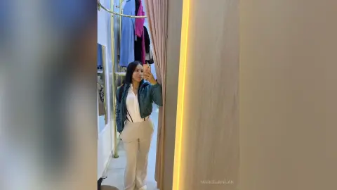 MarcelaMilan takes a mirror selfie wearing a white top, beige pants, and a blue jacket in a dressing room.