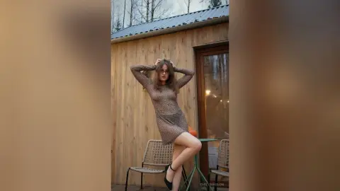 Young woman in a patterned mini dress posing outside a rustic wooden cabin in a serene outdoor setting.