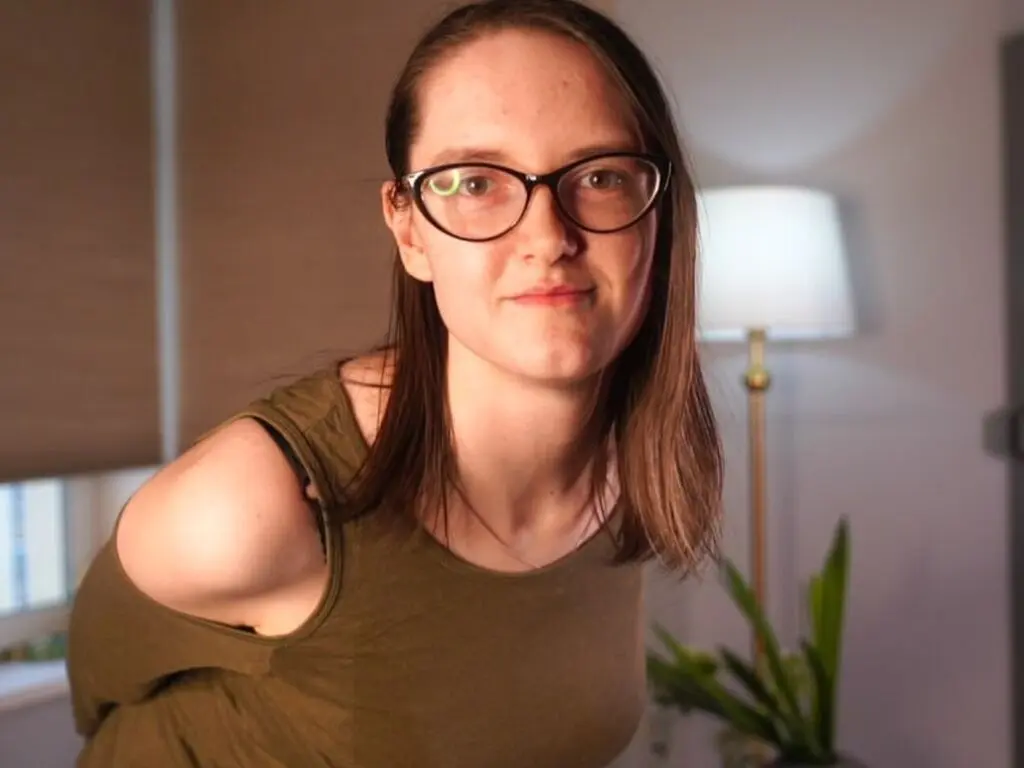 Young woman with glasses in an olive green tank top smiling in a cozy, dimly lit indoor setting.