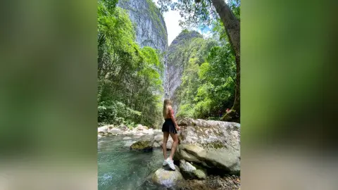 Young woman in a pink top and black skirt standing on rocks by a lush tropical river canyon.