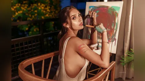 LucyMartin poses in a white tank top while holding a paintbrush near her lips, sitting in a wooden chair next to a colorful painting.