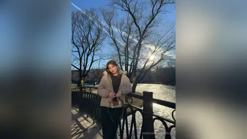 MargaritaHafele stands on a bridge outdoors wearing a tan hooded jacket and dark pants, with bare trees and a bright sky behind her.